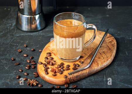Dalgona Kaffee mit Schaum in Glasbecher auf Schneidebrett, Kaffeebohnen und Metallstroh, schwarzer Hintergrund, Nahaufnahme Stockfoto