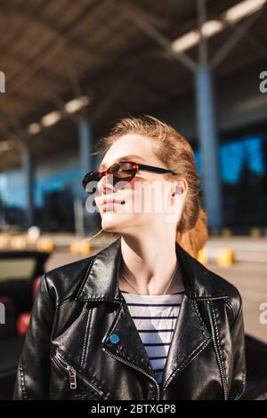 Porträt von schönen lächelnden Mädchen in schwarzer Lederjacke und Sonnenblessen verträumt beiseite mit Flughafen auf Hintergrund zu suchen Stockfoto