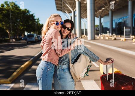 Zwei schöne Mädchen in Sonnenbrille glücklich umarmen einander, während in der Kamera mit roten Koffer und Rucksack auf der Schulter im Freien in der Nähe des Flughafens suchen Stockfoto