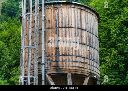 Der hölzerne Wassertank auf dem Hintergrund von grünen Bäumen. Der Tank ist auf einer Holzstruktur mit Rohren und einer Leiter angebracht. Stockfoto