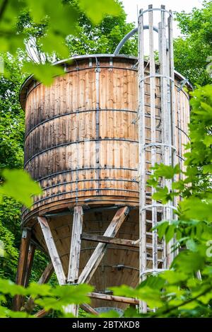 Der hölzerne Wassertank auf dem Hintergrund von grünen Bäumen. Der Tank ist auf einer Holzstruktur mit Rohren und einer Leiter angebracht. Stockfoto