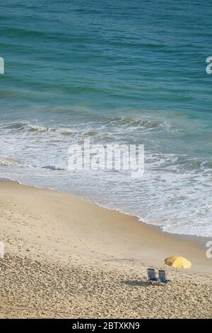 Vertikales Foto eines Strandliegenpaares mit Sonnenschirm am leeren Strand mit den Meereswellen Stockfoto