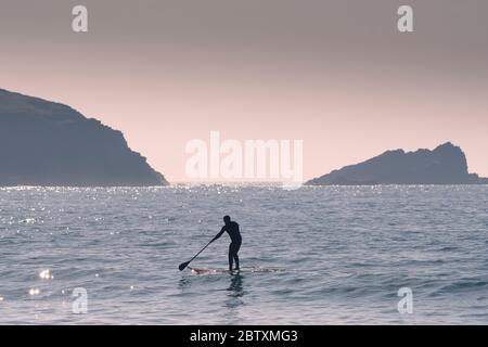 Die Silhouette einer Stand Up Paddle Boarder bei Fistral mit Goose Rock und Pentire Point East im Hintergrund in Newquay in Cornwall. Stockfoto