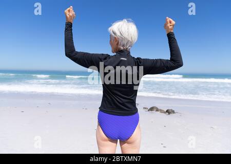 Ältere kaukasische Frau genießt Zeit am Strand an einem sonnigen Tag, stehen und biegen ihren Bizeps mit Meer im Hintergrund Stockfoto