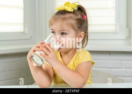 4 Jahre altes süßes Mädchen in einer gelben Bluse trinkt Milch aus einem Glas in der Küche Stockfoto