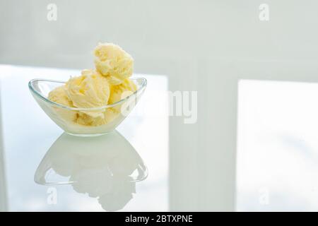 Eine Glasschale mit Vanilleeis steht auf einem hellen Tisch in der Küche. Schüssel, Fenster und Himmel spiegeln sich im Tisch wider Stockfoto