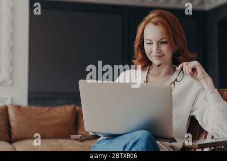 Horizontale Aufnahme von glücklichen geschickten Ingwer Frau Lehrer arbeitet entfernt, prüft Studenten arbeitet über Laptop, hält Brillen, posiert in modernen gemütlichen Wohnung Stockfoto