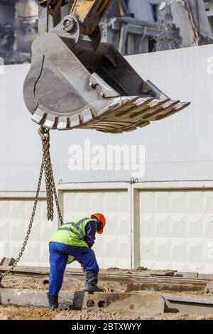 Baggerschaufel heben Metallbalken mit Arbeiter helfen, Baustelle und abgerissen Gebäude auf Hintergrund / Stockfoto