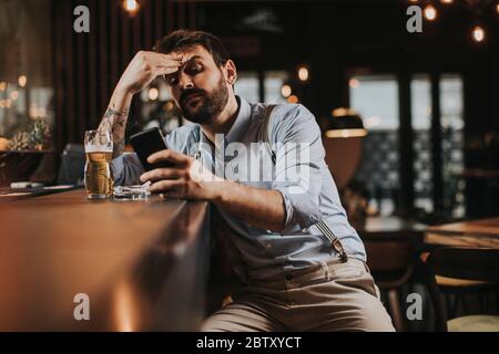 Junger Mann, der Bier trinkt, Zigarette raucht und im Pub Handy benutzt Stockfoto