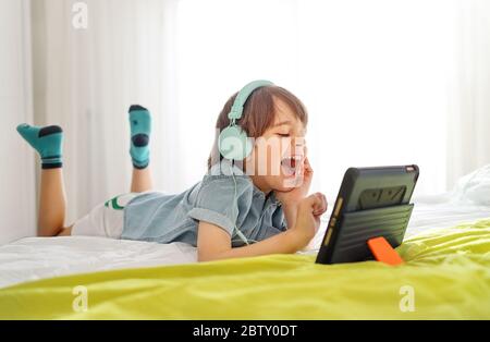 Kleiner lächelnder Junge, der auf dem Bett sitzt und zu Hause auf einem digitalen Tablet spielt. Kind in seinem Schlafzimmer trägt Kopfhörer und mit intelligenten Geräten Spaß Stockfoto