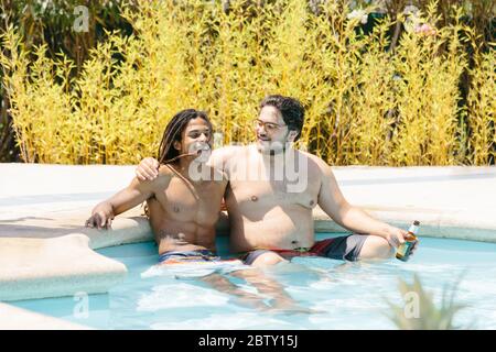 Ein paar multiethnische Männer umarmen sich, während sie mit einer Bierflasche in der Hand in einem Pool sitzen Stockfoto