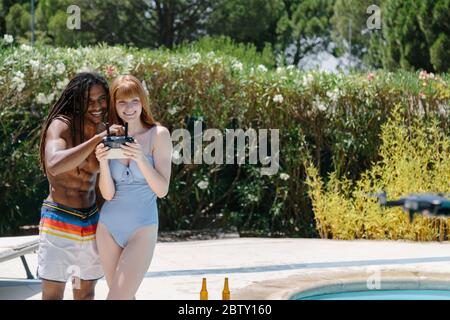 Multikulturelles Paar in Badeanzug, das in einem Pool mit einer Fernbedienung einer Drohne an einem sonnigen Tag steht, umgeben von einem Garten und Gebäuden Stockfoto
