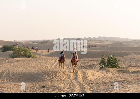 Ein Kamelie (Kamelfahrer) trägt das dekorierte Kamel, während er mit seinen Mobiles auf sam Sanddünen der Thar Wüste mit Touristen während Sonnenuntergang spricht Stockfoto