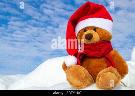 Weihnachtsbär in einem Weihnachtsmann Hut und einem roten Strickschal um den Hals gebunden. Ein Teddybär sitzt in einer Schneewehe auf einem Hintergrund von blauem Himmel. Stockfoto
