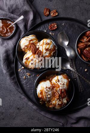 Zwei schwarze Schalen mit Vanille-Eiskugeln mit Karamellsauce und Pekannüssen auf dunklem Marmorgrund. Draufsicht Stockfoto
