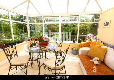 Innenraum eines hellen, sonnendurchfluteten Wintergartens mit Glasfenstern, Tischen, Stühlen und Sofa mit Garten im Hintergrund Stockfoto