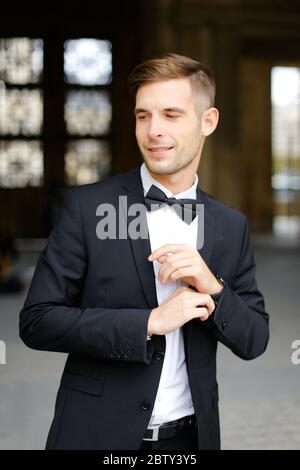 Junger, schöner Mann steht und posiert, trägt schwarzen Anzug und Fliege. Stockfoto