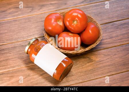 Viele rote Tomaten in einem gewebten Korb mit einem Glas Tomatenmark auf einem Holzbrett Stockfoto