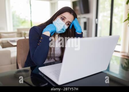 Junge traurige Geschäftsfrau, die von zu Hause aus am Laptop arbeitet, Schutzmaske und Handschuhe trägt. Coronavirus Quarantäne und Geschäftsprobleme Konzept. Stockfoto