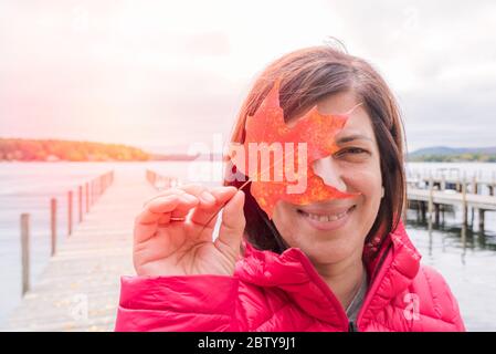 Portrain einer lächelnden Frau, die ein rotes Ahornblatt vor ihrem rechten Auge hält. Im Hintergrund ist ein verlassener Steg auf einem See zu sehen. Stockfoto