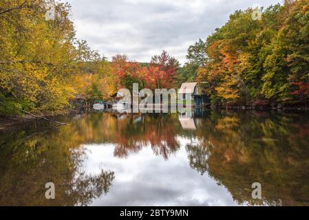 Bootshäuser und Boote entlang eines Flusses mit bewaldeten Ufern an einem bewölkten Herbsttag. Atemberaubende Herbstfarben und Reflektionen im Wasser. Stockfoto