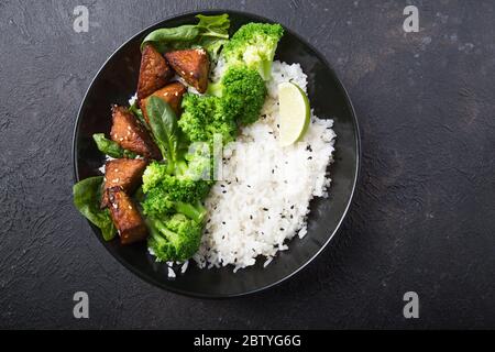 Vegane Teryaki Tempeh oder tempe buddha Schüssel mit Reis, gedämpftem Brokkoli, Spinat und Limette auf schwarzem Hintergrund. Gesunde Ernährung Stockfoto