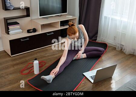 Rotschopf Mädchen in Sportbekleidung Stretching zu Hause, sitzt vor einem Laptop, Rahmen von oben. Stockfoto