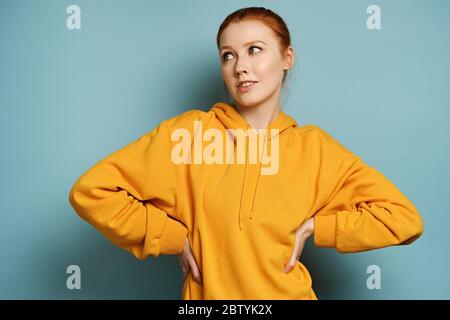 Ein rothaariges Mädchen in einem gelben Sweatshirt steht auf blauem Hintergrund, schaut zur Seite und legt seine Hände auf den Gürtel. Stockfoto