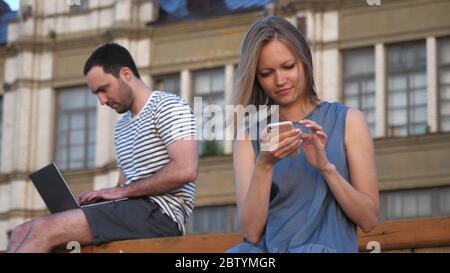 Mann mit Laptop und Frau mit Smartphone sitzen auf einem Bank o Stockfoto