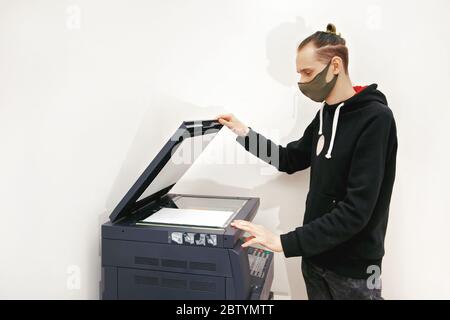 Der junge Mann in einer Schutzmaske macht Kopien von Dokumenten auf einer Kopiermaschine im Büro Stockfoto