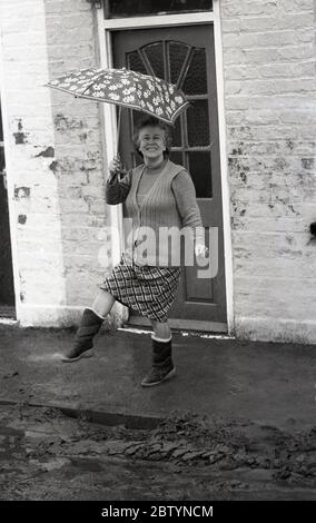 Vor ihrem kleinen Reihenhaus, mit nassem Schlamm in der Rinne, eine Dame mittleren Alters hält einen Regenschirm und macht ein wenig Tanz, vielleicht in Erleichterung, dass das Hochwasser nicht in ihr Haus, England, Großbritannien, 1987. Stockfoto