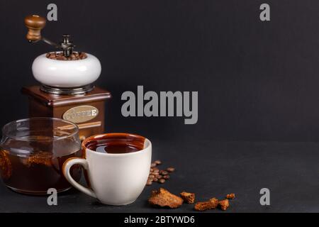 Zubereitung Chaga Pilze Kaffee. Weiß-braune Keramikbecher und Glasgefäß mit Chaga-Getränk und Kaffeemühle auf schwarzem Hintergrund. Trendy gesund beve Stockfoto