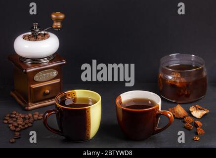 Zubereitung Chaga Pilze Kaffee. Zwei zweifarbige Keramikbecher, Glasgefäß mit Chaga-Getränk, Kaffeemühle auf schwarzem Hintergrund. Trendige gesunde Bevera Stockfoto