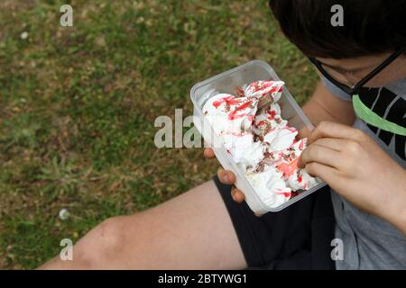 Junge, die Eis aus der Wanne mit Plastiklöffel und schmuddeligen Händen essen, UK, Sommer 2020 Stockfoto