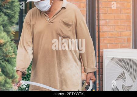 Ein alter Mann mit Quarantäne trägt eine Gesichtsmaske, um die Ausbreitung des Corona-Virus (Covid-19) zu verhindern, das Pflanzen für die Bewegung im Garten zu Hause bewässert. Nein Stockfoto