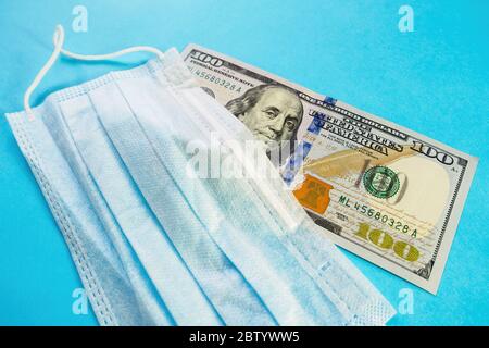 Gesichtsmaske liegt auf einer Banknote von 100 Dollar auf blauem Hintergrund. Medizinisches Konzept zur Preissteigerung von Masken, Schutz vor Coronavirus, Grippe und Allergie. Whirlpool kopieren Stockfoto