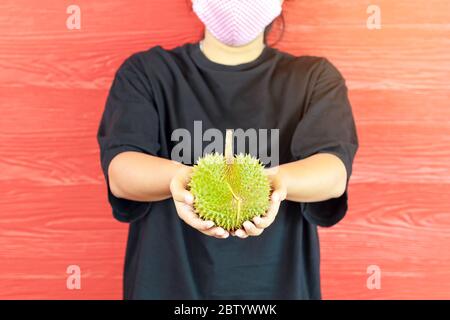 Junge Frau trägt eine Schutzmaske, um eine Infektion des Corona-Virus (Covid-19) in einem schwarzen Hemd mit einem reifen Durian zu verhindern, bereit, mit dem roten w zu essen Stockfoto