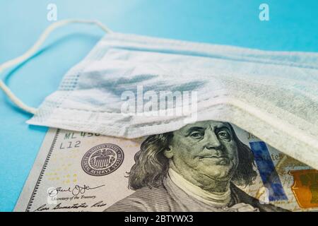 Medizinische Maske und Banknote von 100 Dollar auf blauem Hintergrund. Konzept von Defizit, Spekulation und ausverkauften Gesichtsmasken. Selektiver Fokus, Nahaufnahme Stockfoto