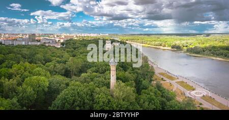 Stadtpanorama öffentlicher Park mit großer Grünfläche in der Nähe des Flusses ein alter Leuchtturm in den Bäumen ein blauer Himmel mit Cumuluswolken auf der Landschaft. Luftaufnahme Stockfoto