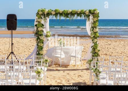 Romantische Trauung am Meer, Strand an einem sonnigen Tag. Ein Holzbogen mit frischen Blumen. Weiße Sessel im Sand. Stockfoto