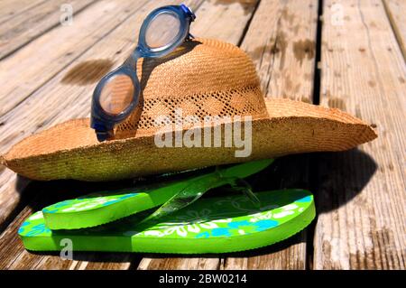 Das Notwendige zum Schwimmen lag auf einer rustikalen Holzterrasse. Ein Strohhut, eine Brille und grüne Flip-Flops liegen in einem Haufen. Stockfoto