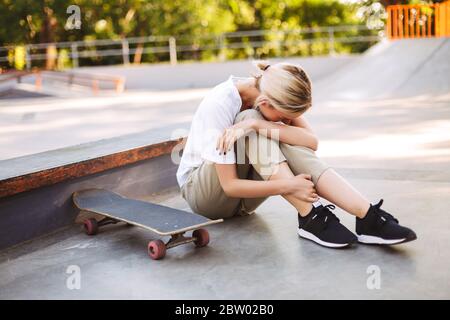 Junge Skater Mädchen hält ihr schmerzhaftes Bein und weint mit Skateboard in der Nähe Skatepark isoliert Stockfoto