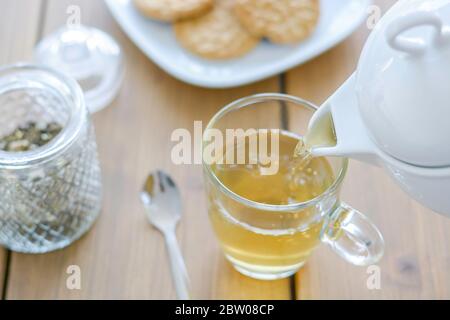 Weiße Teekanne, Glaskuette, loser Tee und Kekse im Hintergrund. Tee eingießen. Holztisch. Stockfoto