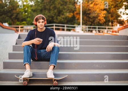 Junge Skater in orange Kopfhörer halten Handy in den Händen glücklich in der Kamera auf Treppen mit Skateboard in modernen Skatepark suchen Stockfoto