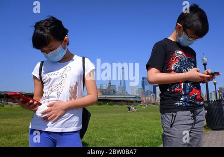 Junge asiatische Mädchen und Jungen tragen Maske Spiele auf dem Handy im Liberty State Park während des Covid-19 Coronavirus Ausbruch im Liberty State Park mit der Skyline von Lower Manhattan New York City Financial District im Hintergrund. New Jersey.USA Stockfoto