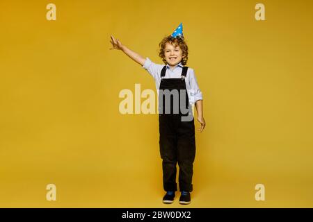 Stilvolle rothaarige kleine Junge in Overalls und Geburtstagsmütze posiert auf dem gelben Hintergrund, isoliert. Kindermode Stockfoto