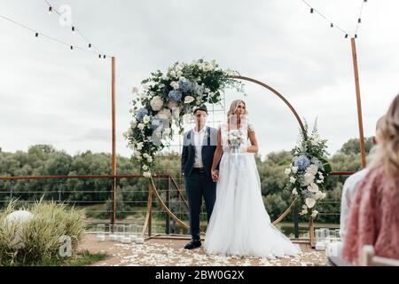 Glücklich frisch vermählten, wo Braut höher ist als Bräutigam am Hintergrund der Hochzeit Bogen stehen. Hochzeitszeremonie Stockfoto