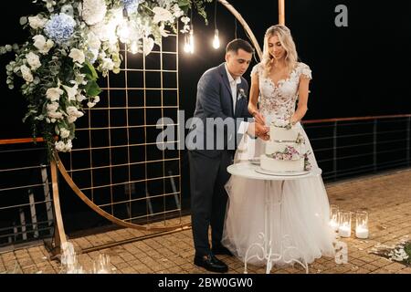 Glückliche Braut und Bräutigam stehen in der Nähe der Hochzeit Bogen und schneiden ihre Hochzeitstorte Stockfoto
