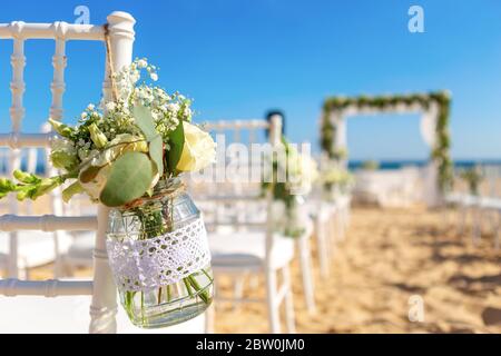 Luxuriöse Hochzeitszeremonie am Meer, Strand. Weiße Stühle mit einem schönen Blumenstrauß in einem Glas, das darauf hängt, dekoriert. Stockfoto