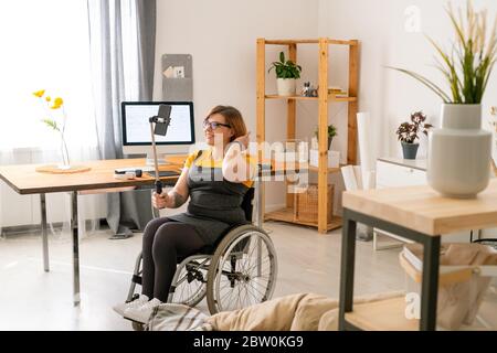 Fröhliche behinderte junge Bloggerin in Brille im Rollstuhl sitzen und Video-Streaming zu Hause Stockfoto